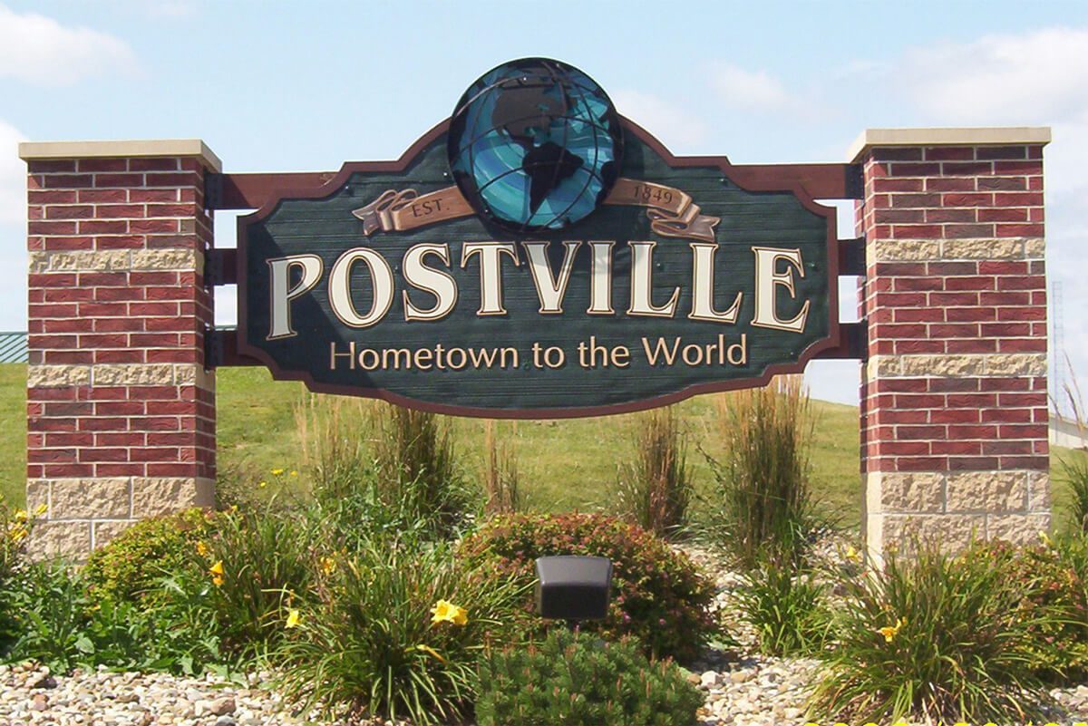 Postville sign saying "Postville Hometown to the World Est 1849" with flowers underneath.