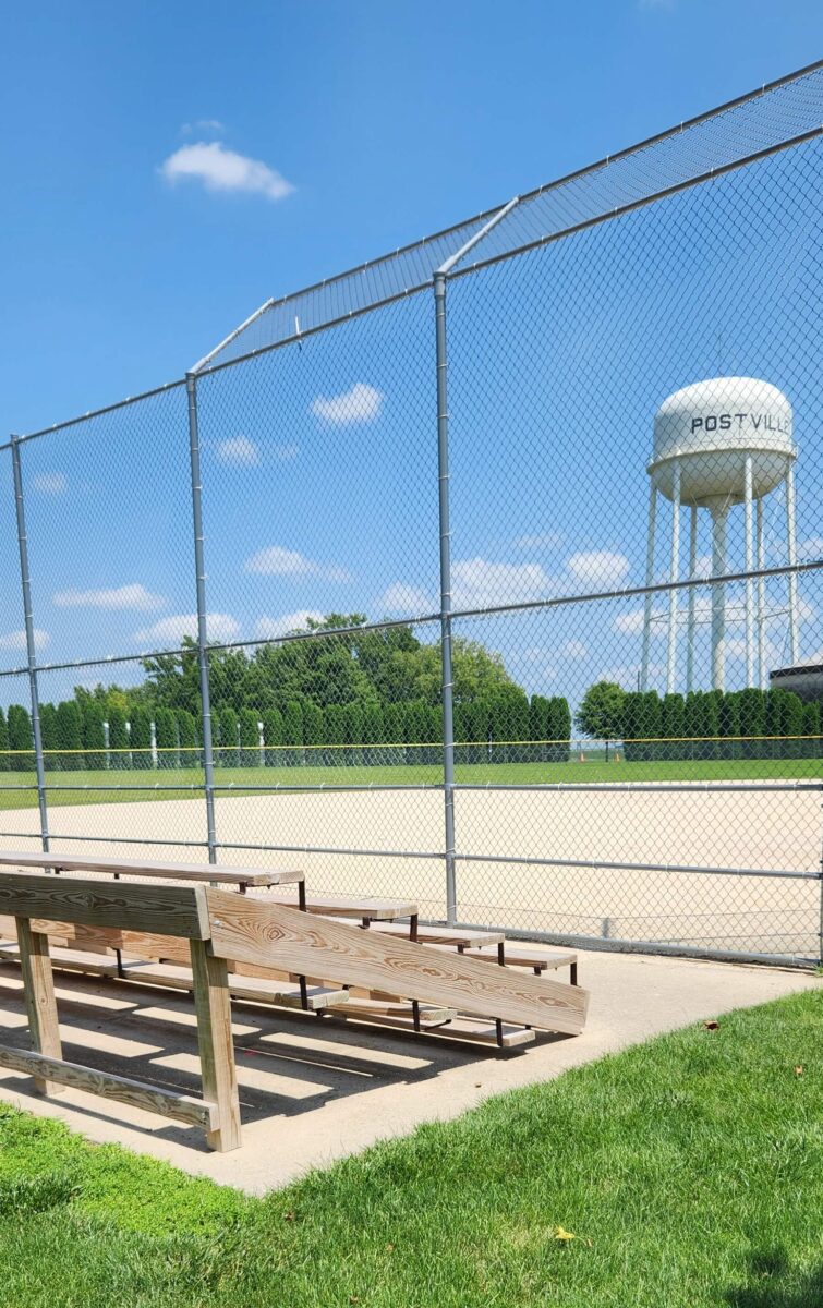 A baseball field with a wooden bench in the foreground. The field is enclosed by a chain-link fence. In the background, there is a water tower labeled “POSTVILLE” and a row of trees under a blue sky with a few scattered clouds.