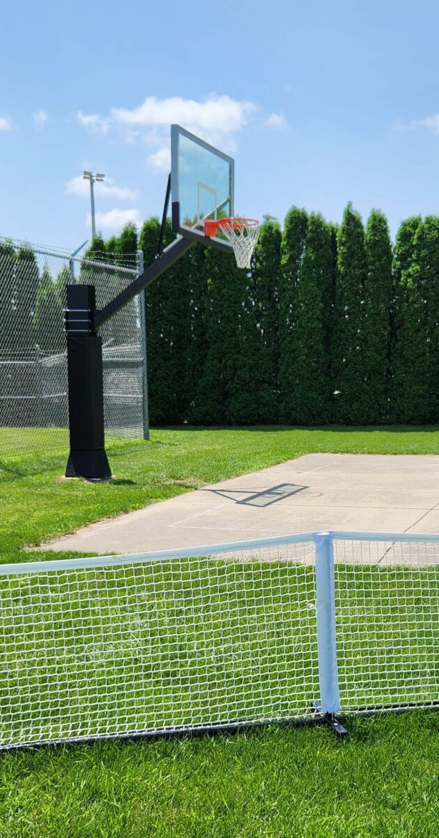 A basketball hoop stands on a concrete court, surrounded by a chain-link fence. Tall, dense trees form a green backdrop. In the foreground, a white mesh barrier sits on the grass. The sky is clear and blue.