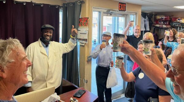 A group of cheerful people inside a small shop or café raise their coffee cups in a toast. A smiling man, dressed in white with a cap, stands behind a counter, giving a thumbs up. The atmosphere appears joyful and celebratory.