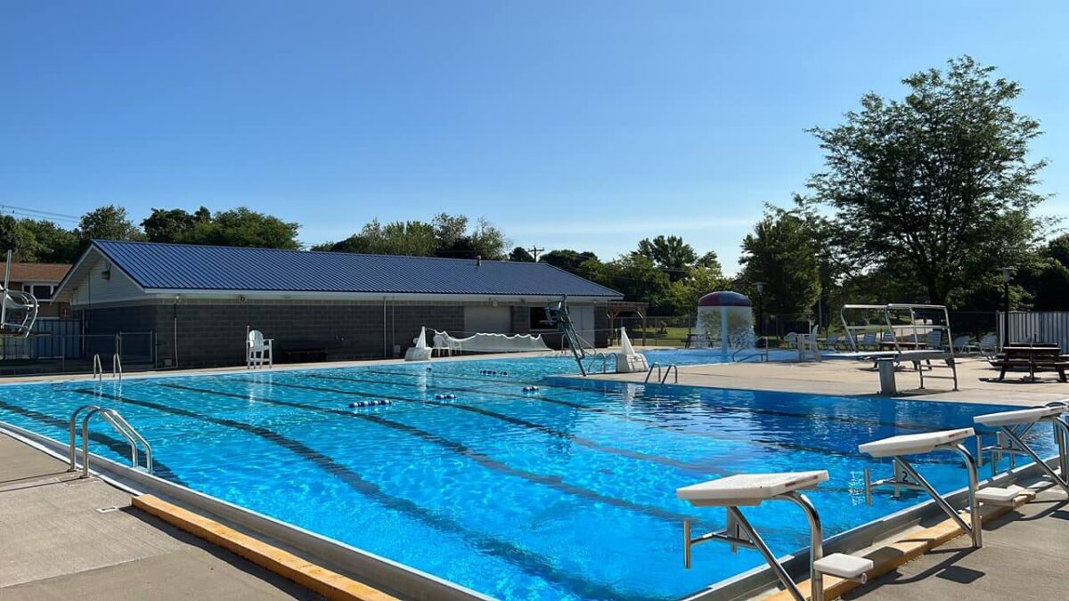 An outdoor swimming pool is shown on a clear, sunny day. The pool features swim lanes, diving boards, and a lifeguard stand. A building with a blue roof is in the background, along with trees and shaded areas. Poolside chairs and a slide are also visible.