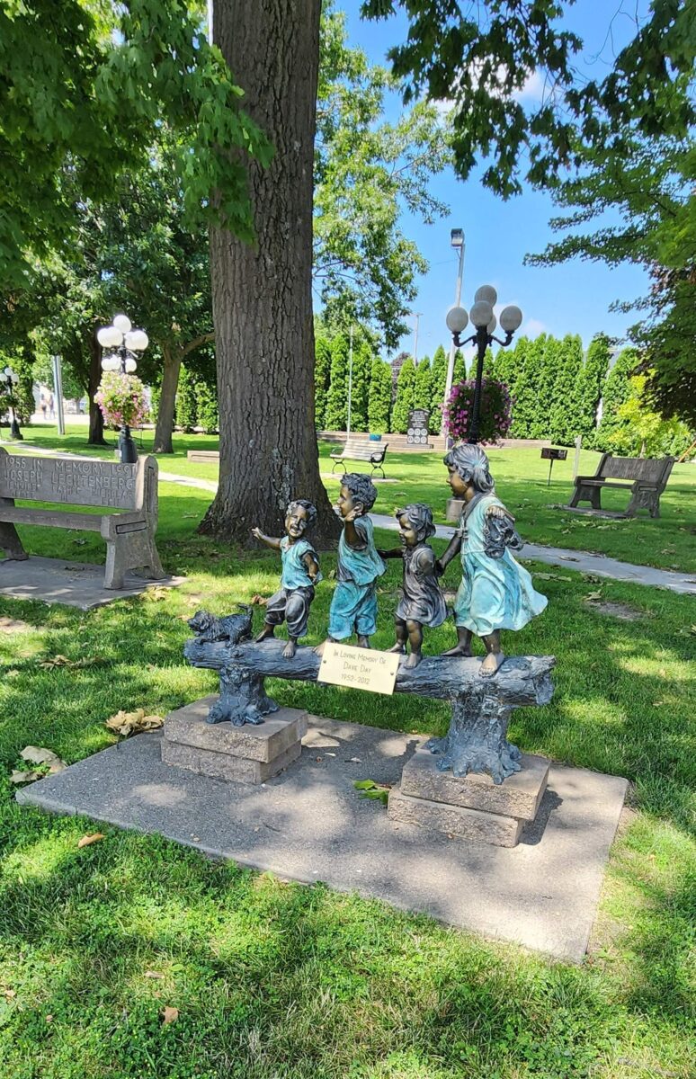 A bronze sculpture in a park depicts four children playfully walking on a tree branch. The sculpture is surrounded by green grass and trees, with a few park benches and lampposts in the background. A yellow sign is placed in front of the sculpture.