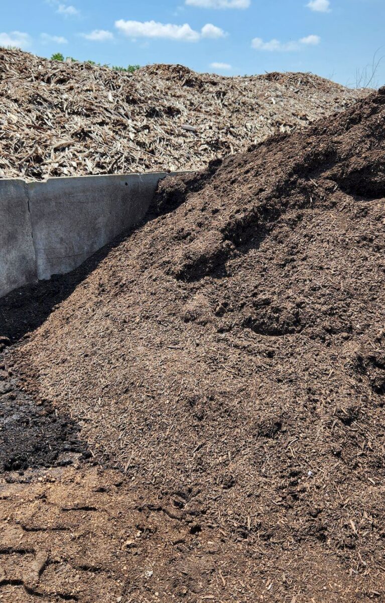 A large pile of dark brown mulch is shown in an outdoor setting under a partly cloudy sky. The mulch pile is surrounded by other heaps of organic material and a concrete wall to the left. The scene appears to be part of a landscaping or composting area.