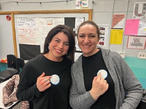 Two women standing side by side in a classroom, both smiling and pointing at circular badges pinned to their black tops. The classroom has posters, charts, and a whiteboard with writing. The women have light skin tones and dark hair.