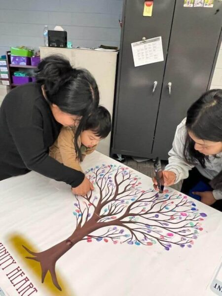 Three individuals, an adult and two children, are placing colorful fingerprints on the branches of a tree design on a large white sheet of paper. The scene appears to be in a classroom with a metal cabinet and various supplies in the background.