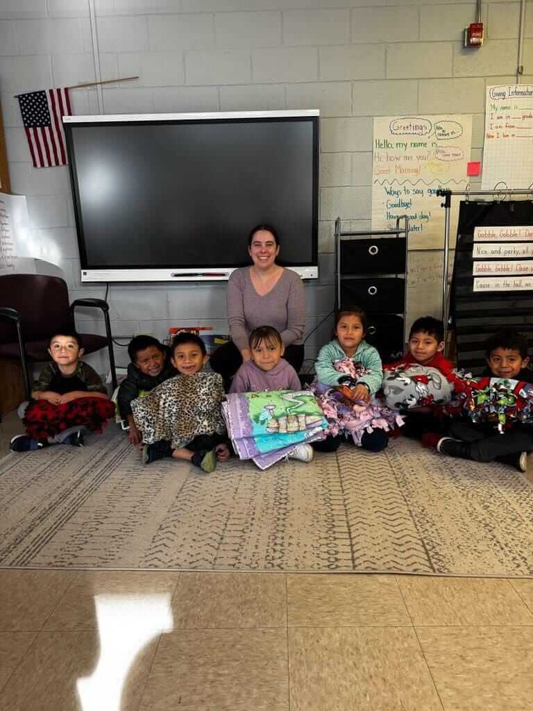 A woman sits on the floor with a group of children in a classroom. Behind them is a large screen and an American flag. The children are holding up colorful blankets, and there are posters with educational content on the walls.