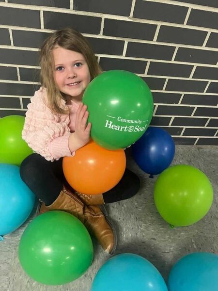 A young girl with blonde hair, wearing a pink sweater and brown boots, sits on the floor holding balloons of various colors. She smiles at the camera while surrounded by more balloons. One green balloon she holds has "Heart & Soul Community" printed on it.