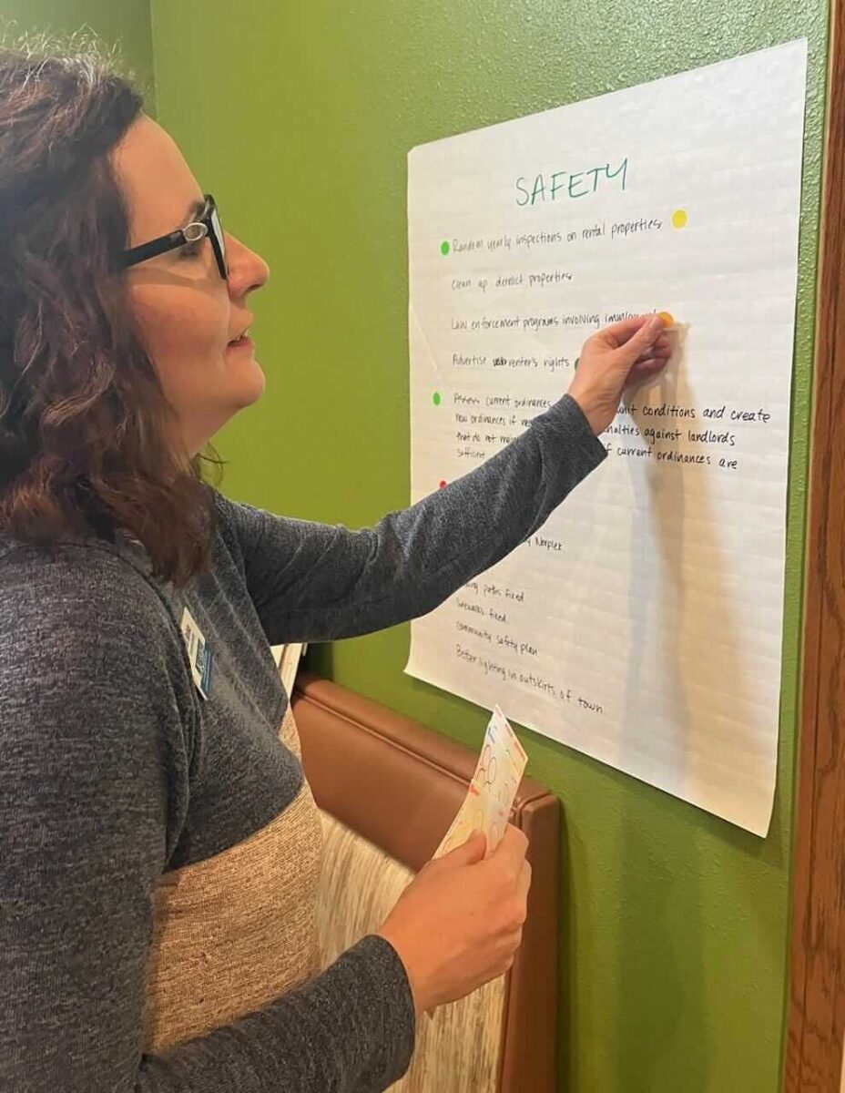 A woman with glasses and curly hair is pointing to a handwritten poster on a wall labeled "SAFETY." The poster outlines various safety protocols and precautions. She is wearing a grey and beige sweater and appears to be engaged in explaining the details.