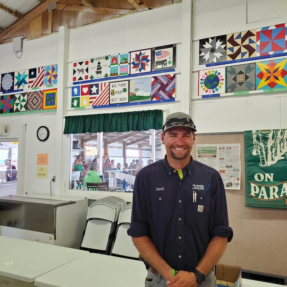 A smiling man wearing a navy blue shirt, sunglasses on his cap, and a name tag stands in front of colorful quilt designs displayed on the wall in a community room. There are folding chairs, tables, and a green banner in the background.