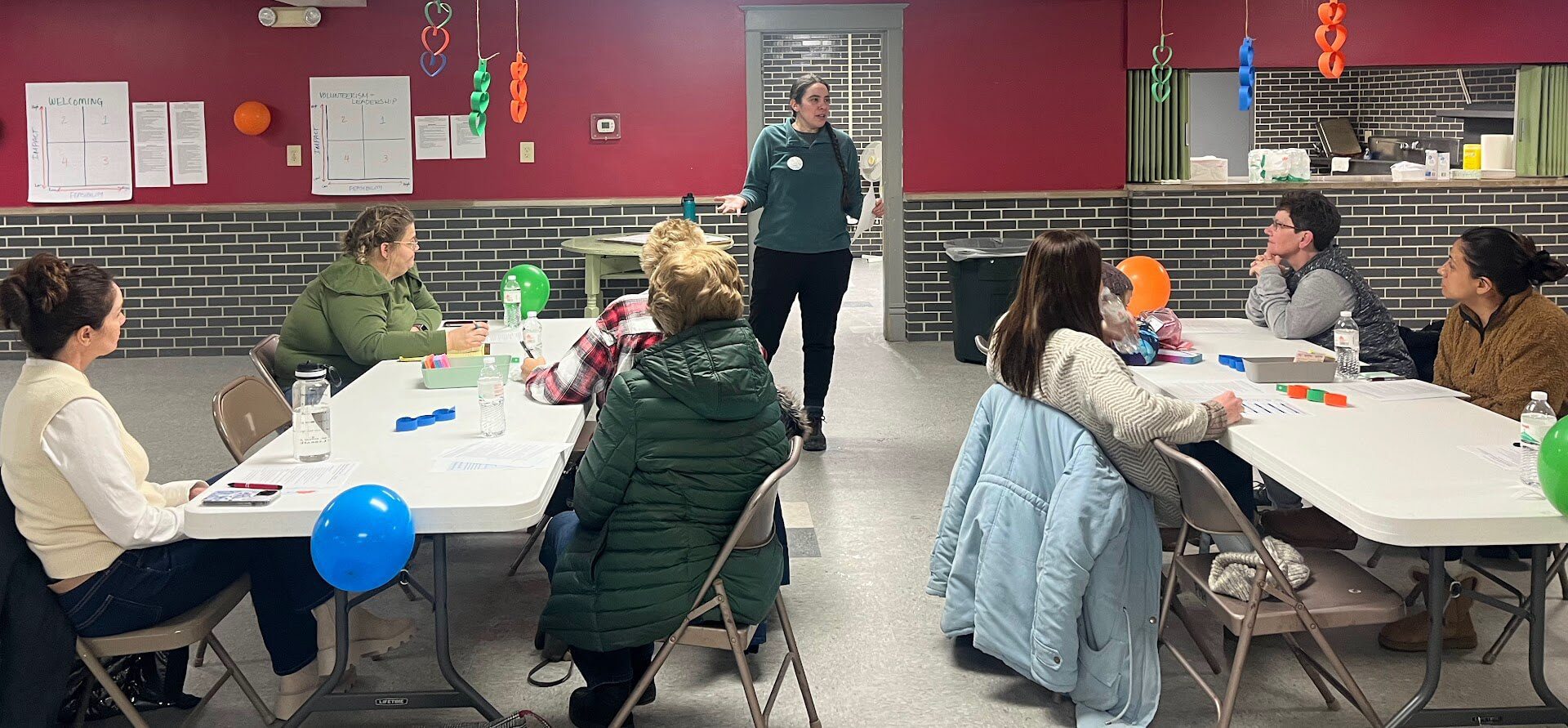 A group of people sit at tables in a community center, listening to a person stand and speak at the front of the room. The tables have balloons and papers on them, and the walls are decorated with colorful streamers. The atmosphere appears to be casual and attentive.