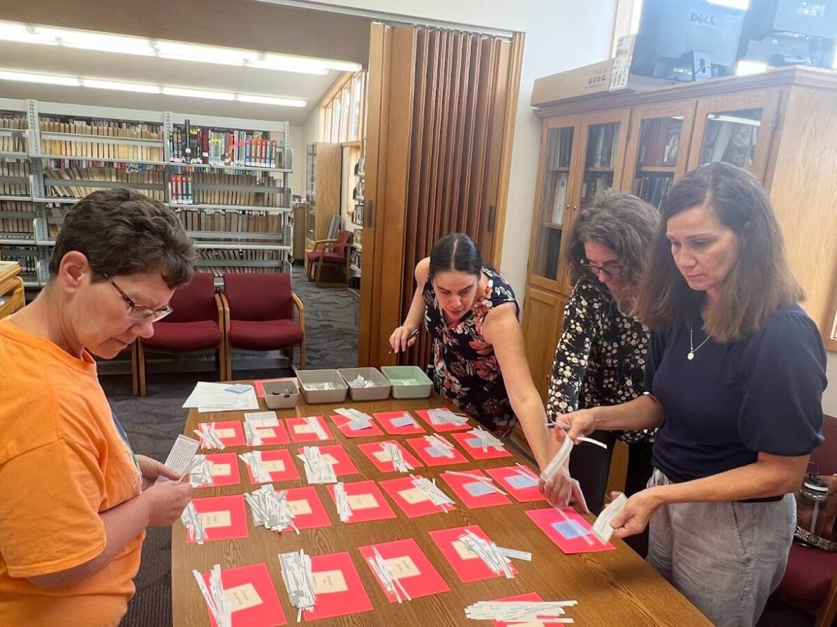 Four people are gathered around a table in a library, engaged in organizing name tags or cards. The table is covered with red paper that has text, and several containers are nearby. Bookshelves filled with books are visible in the background.