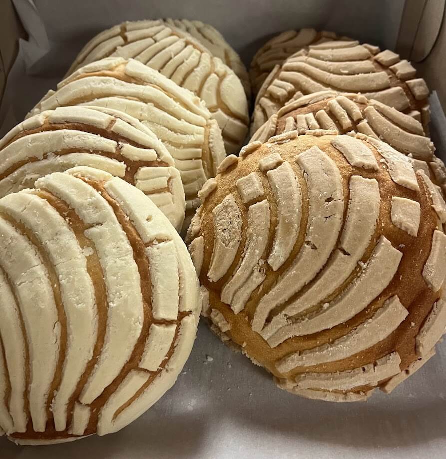 A box filled with conchas, a traditional Mexican sweet bread. The conchas have a distinctive shell-like pattern on top, with some appearing white and others brown. The bread has a soft, fluffy texture, visible through the light crust on top.