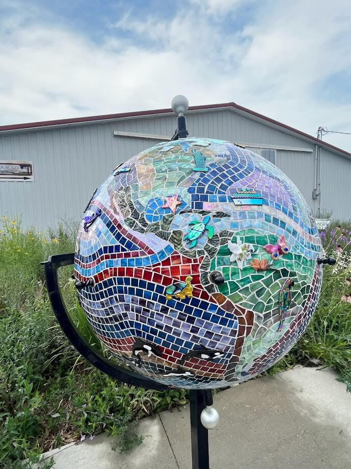 A colorful mosaic sculpture of a globe stands outdoors in front of a gray building. The globe is adorned with various tiles and small 3D elements like flowers, birds, and animals, creating a vivid, artistic representation of Earth. The sky is partly cloudy.