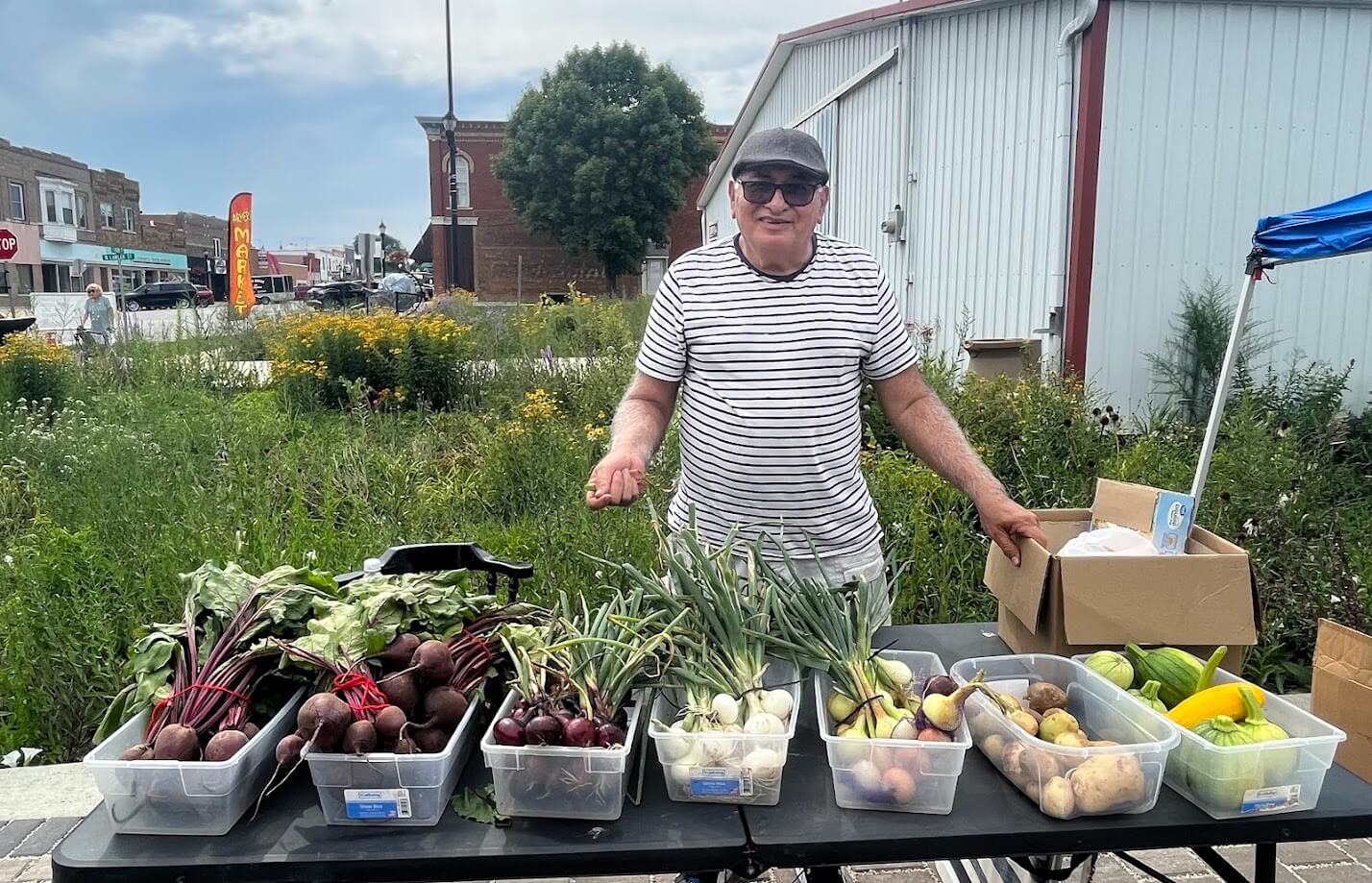 A person in a striped shirt and hat is standing behind a table at an outdoor market. The table has bins filled with various fresh vegetables including beets, onions, potatoes, and peppers. Buildings and greenery are visible in the background.