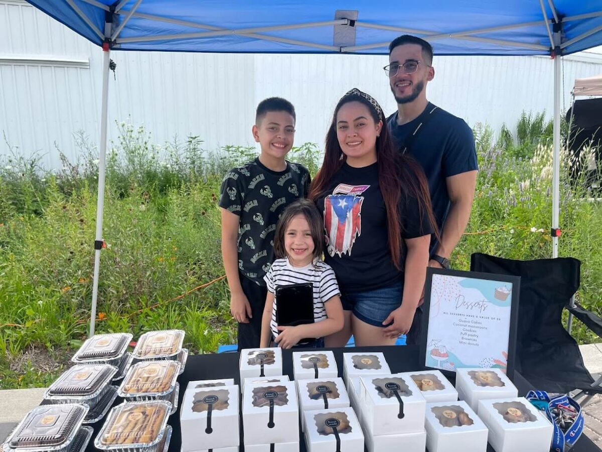 A family of four smiles while standing behind a table displaying neatly organized baked goods under a blue canopy at an outdoor market. The table features various packaged treats and a colorful menu. The background shows foliage and a white building.