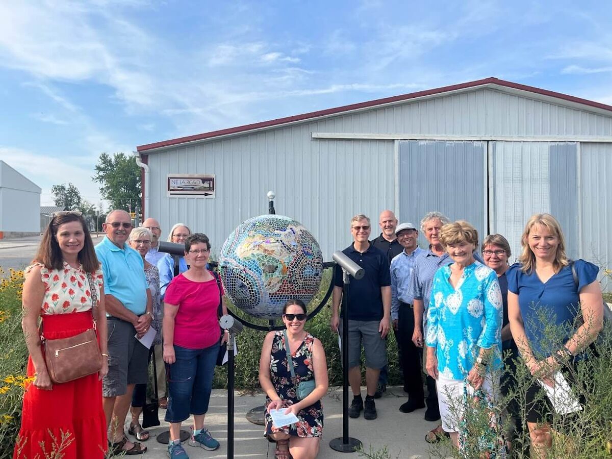 A group of 14 people of various ages are standing and sitting around a large, artistic globe sculpture outside a light grey building with a red roof. The sky is clear and sunny. Some are smiling at the camera while others look towards the globe.