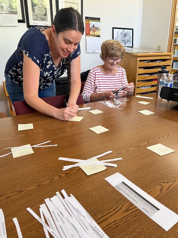 Two women collaborate at a table, organizing paper strips and sticky notes. The younger woman with dark hair leans over to write on a note, while the older woman with glasses and short hair cuts paper strips. Both are engaged in focused work.