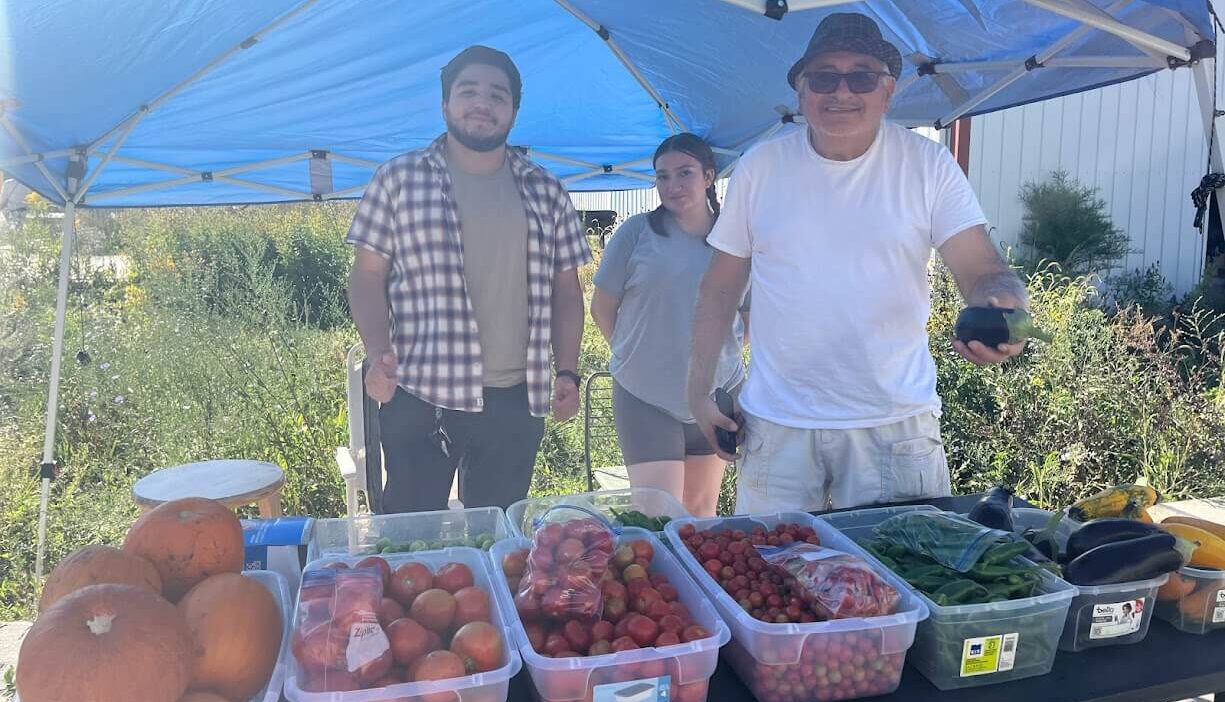 Three people stand under a blue canopy at a market stall featuring various fresh produce, including pumpkins, tomatoes, and greens. The person on the right is holding a dark object, possibly a phone. The backdrop includes greenery and a white building.