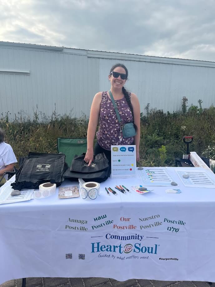 A woman wearing a purple patterned tank top and sunglasses stands behind a table covered with a white tablecloth that reads "Community Heart & Soul." The table displays informational materials and small items. A white building and greenery are in the background.