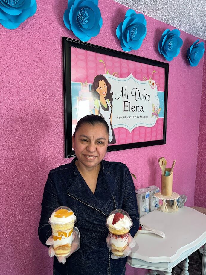 A woman smiling and holding two dessert parfaits in front of a pink wall with blue flower decorations. Behind her is a framed sign that reads, "Mi Dulce Elena," with an illustration of a woman and decorative elements.